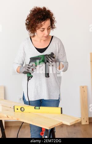 Caucasian woman with curly hair holds a drill in her hands, ready to make home improvements. She is standing in front of a workbench. She wears a gray Stock Photo