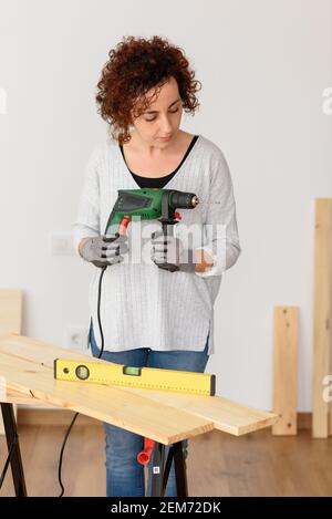 Caucasian woman with curly hair holds a drill in her hands, ready to make home improvements. She is standing in front of a workbench. She wears a gray Stock Photo