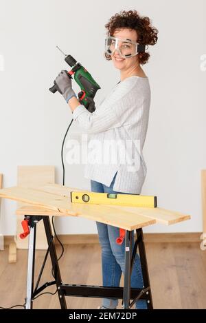 Caucasian woman with curly hair holds a drill in her hands, ready to make home improvements. She is standing in front of a workbench. She wears a gray Stock Photo