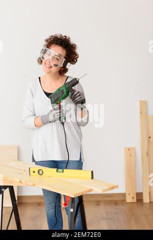 Caucasian woman with curly hair holds a drill in her hands, ready to make home improvements. She is standing in front of a workbench. She wears a gray Stock Photo