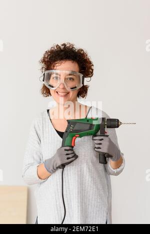 Caucasian woman with curly hair holds a drill in her hands, ready to make home improvements. She is standing in front of a workbench. She wears a gray Stock Photo