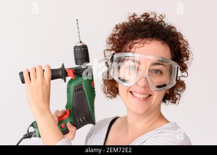 Caucasian woman with curly hair holds a drill in her hands, ready to make home improvements. She is standing in front of a workbench. She wears a gray Stock Photo