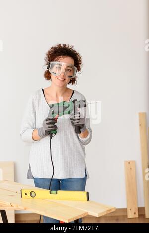 Caucasian woman with curly hair holds a drill in her hands, ready to make home improvements. She is standing in front of a workbench. She wears a gray Stock Photo
