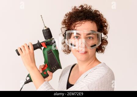 Caucasian woman with curly hair holds a drill in her hands, ready to make home improvements. She is standing in front of a workbench. She wears a gray Stock Photo