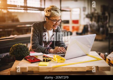 Cheerful smiling beautiful middle-aged female engineer looking on a laptop on the desk in the sunny workshop. Stock Photo