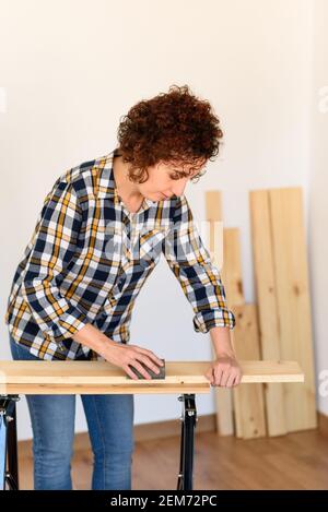 Girl doing DIY and handicraft tasks, dressed in a plaid shirt and jeans. She is Caucasian and has curly hair. She is sanding some wood on a workbench Stock Photo