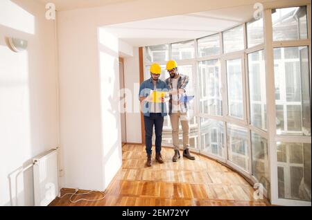 Two handsome young engineers with safety hats are explaining ideas to each other by pointing out on blueprints on their folders near windows of very b Stock Photo