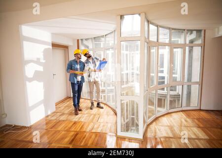 Two charming young architects with yellow safety hats are checking progress on their projects comparing interior with blueprints near a window in a ve Stock Photo