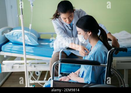 Asian woman doctor using a stethoscope to check the heart rhythm of a female patient wearing face mask while sitting on a wheelchair at hospital Stock Photo