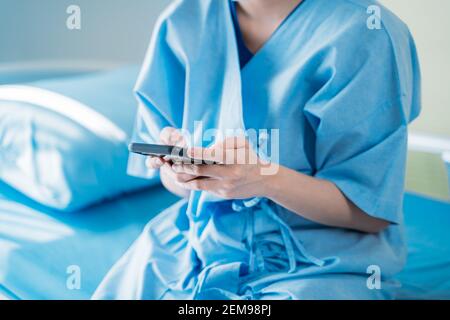 Closeup of hands of young female patient using smartphone to text and browse internet while sitting on hospital room bed wearing scrub uniform Stock Photo