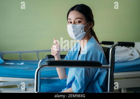 Portrait of a smiling Asian young female patient with surgical mask sitting on wheelchair in hospital room showing thumbs up gesture and sign to camera Stock Photo