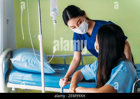 Young nurse wearing surgical mask in conversation with and comforting depressed Asian patient sitting in wheelchair in hospital ward room Stock Photo