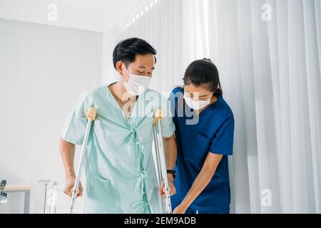 Young female Asian physical therapist in uniform helping man walk using crutches walk in hospital passage wearing face mask Stock Photo