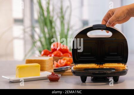 Hand opening the lid of a sandwich making machine with delicious sandwiches next to tomato sauce and butter. Selective focus. Homemade food concept. Stock Photo