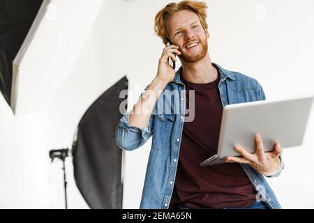 Smiling male photographer using laptop computer while sitting at the studio, talking on mobile phone Stock Photo