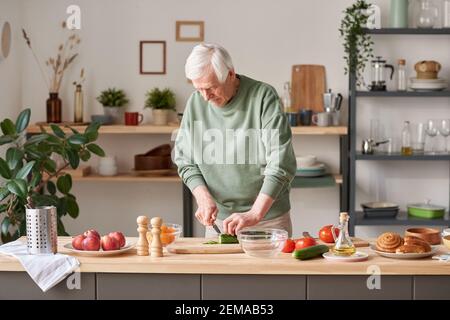 Senior man standing at the kitchen table and cutting vegetables on cutting board in the kitchen Stock Photo
