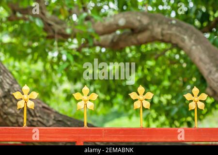 Orange steel fence decorated with wrought iron golden flowers isolated on blurred branches of mango tree backgrounds. Stock Photo