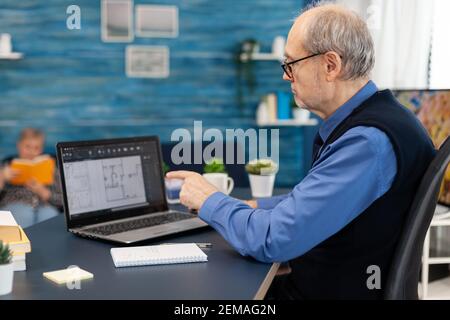 Senior man working with laptop to make home plan. Experienced construction engineer designer looking at house blueprints on laptop computer from home office. Stock Photo