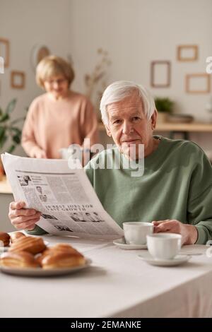 Portrait of senior man having breakfast and reading newspaper at the table with his wife in the background Stock Photo