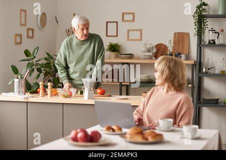 Senior man preparing food and talking to his wife while she working on laptop at the table in the kitchen Stock Photo