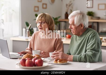 Senior couple sitting at the table with cups of tea and using laptop computer watching something while sitting in the kitchen Stock Photo