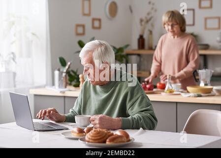 Senior man sitting at the table and typing on laptop he working online at home while his wife cooking in the background Stock Photo