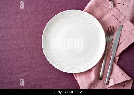 Clean empty white plate, fork and knife on bright blueberry red linen tablecloth on table, mockup Stock Photo