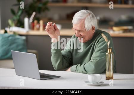 Happy senior man sitting at the table and looking at computer monitor talking online at home Stock Photo