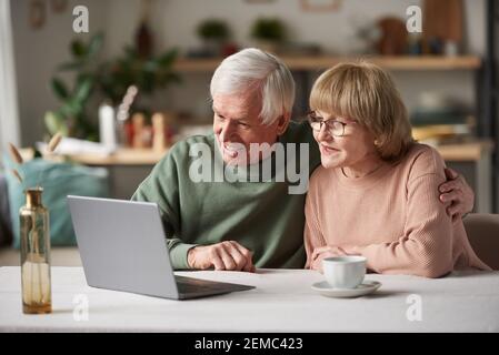 Senior couple sitting at the table in front of laptop looking at monitor and talking online at home Stock Photo