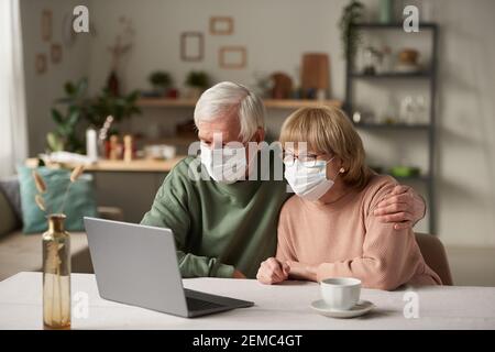 Senior couple in protective masks sitting at the table in front of laptop and talking online with family using laptop during pandemic Stock Photo