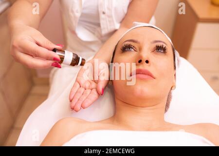 Masseuse applying massage oil on her hands before starting a facial massage on a middle aged woman, facial massage concept Stock Photo