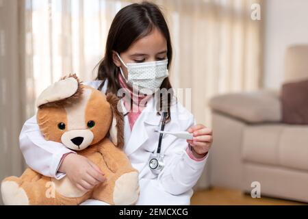 Little child in doctor's uniform taking the temperature of a teddy bear. She is at home. Concept of health and wellness. Space for text. Stock Photo