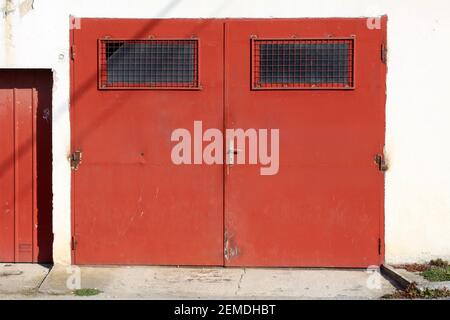 Strong old dark red metal garage doors with rusted hinges and windows covered with metal protection net mounted on dilapidated white garage building Stock Photo