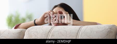 Woman hid behind sofa and peeks out her fingers through the binoculars on her face Stock Photo