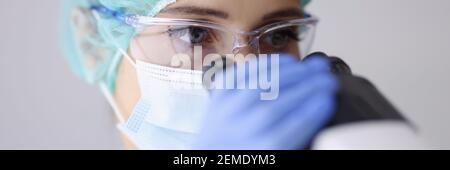 Woman doctor laboratory assistant in goggles and mask looks through microscope. Stock Photo