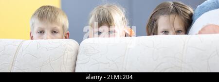 Boy and two girls hide behind sofa and look out. Stock Photo
