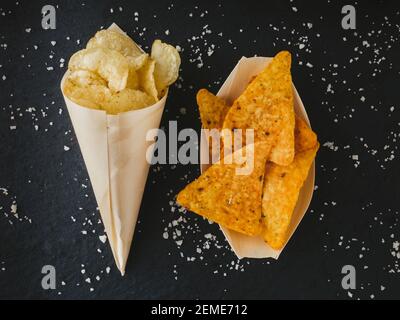 food background. Chips for appetizer. Yellow triangle shaped tortilla chips and potatoes chips on black. Fast salty food for snack or party. Tasty nac Stock Photo
