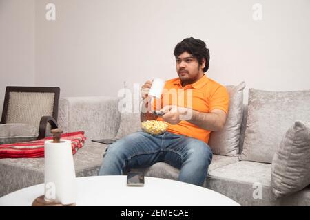Asian Indian man gaming, streaming and watching TV alone in living room on a grey sofa and white table. Stock Photo