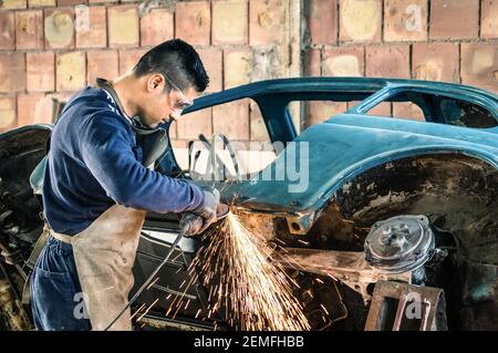 Young man mechanical worker repairing an old vintage car body in messy garage - Safety at work with protection wear Stock Photo