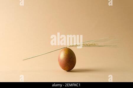 A brown natural chicken egg stands on a beige background. An ear of wheat lies on the egg. Minimalism, monochrome, concept easter background with copy Stock Photo