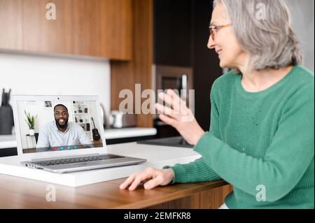 Senior online tutor, trainer, coworker is talking via video connection with an African man on the laptop screen. Mature woman is using computer app for video meeting with biracial colleague, employee Stock Photo
