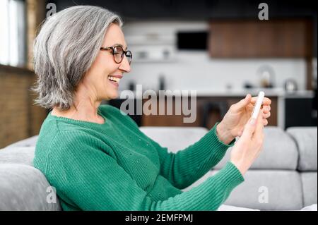 Smiling middle-aged woman with a smartphone sitting on the sofa at home, senior lady has pleasant online chatting on a phone, scrolling news feed, spends time in social networks Stock Photo