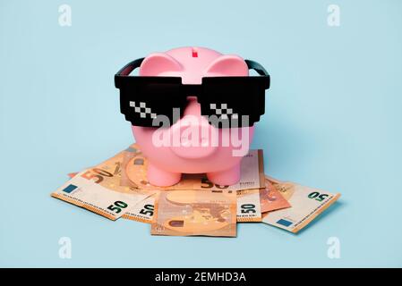 Pink piggy money bank with black sunglasses and euro bills on blue background Stock Photo