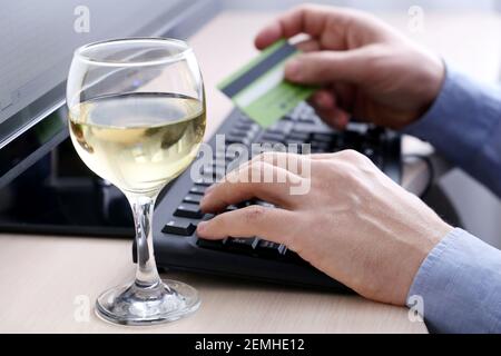 Man holding credit bank card types on PC keyboard, defocused view through glass of white wine. Concept of online shopping under the alcohol Stock Photo