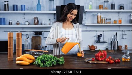 Smiling pregnant woman pouring orange juice near organic vegetables in kitchen Stock Photo