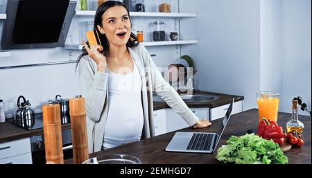 Excited pregnant woman holding credit card near laptop and food in kitchen Stock Photo
