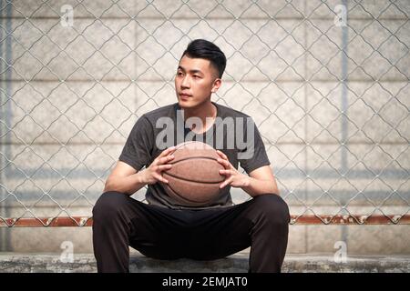 young asian basketball player holding a ball sitting resting at courtside Stock Photo