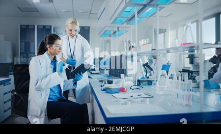 Modern Medical Research Laboratory: Two Female Scientists Working Using Digital Tablet, Analysing Biochemicals Samples. Scientific Lab for Medicine Stock Photo