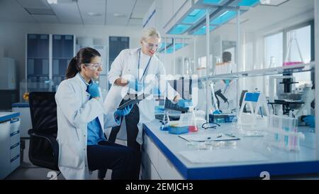 Modern Medical Research Laboratory: Two Female Scientists Working Using Digital Tablet, Analysing Biochemicals Samples. Scientific Lab for Medicine Stock Photo