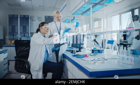 Modern Medical Research Laboratory: Two Female Scientists Working Using Digital Tablet, Analysing Biochemicals Samples. Scientific Lab for Medicine Stock Photo